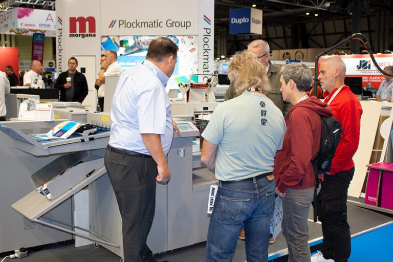 Visitors to The Print Show 2023 gathered around a finishing machine from Morgana whilst a sales man provides a demo