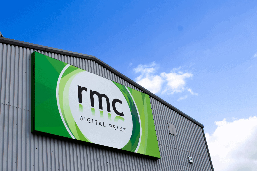 An image of the RMC Digital Print's signage on its warehouse. A green background with a white circle with the letters 'R M C' inside in black font' The sign is attached to a grey metal building