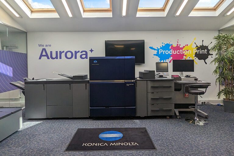Aurora's showroom featuring a Konica Minolta digital printing press in the centre, and a white painted wall behind with the Aurora logo in purple letters and a grey floor with a black rectangle rug showing the Konica Minolta logo
