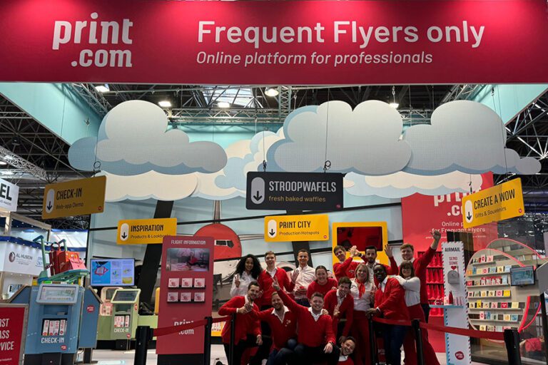The Print.com team wearing magenta, in front of their flight-style themed stand at drupa