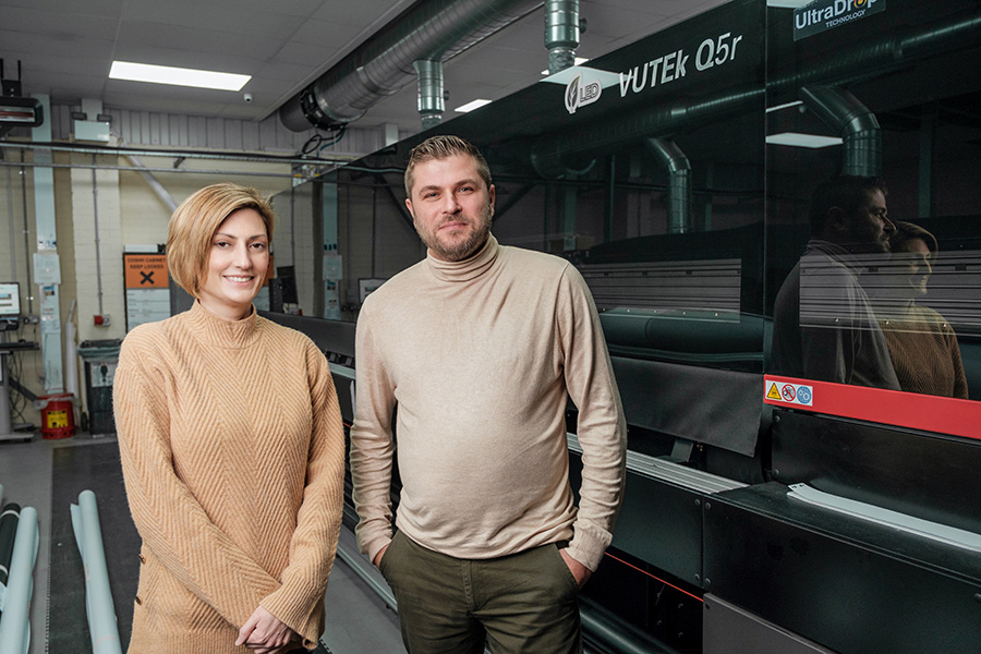 Nicole Spencer, wearing a beige knitted turtle neck dress to the left of other director Alex in front of a large-format printer with a black chassis