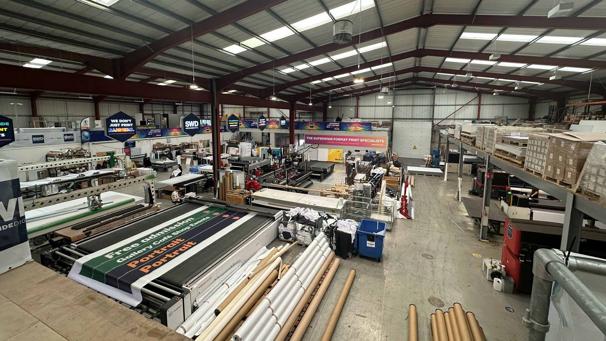 An ariel view of inside a wide-format print warehouse, featuring large wide-format printers, rolls of media, and concrete floor and a hangar-style slanted roof