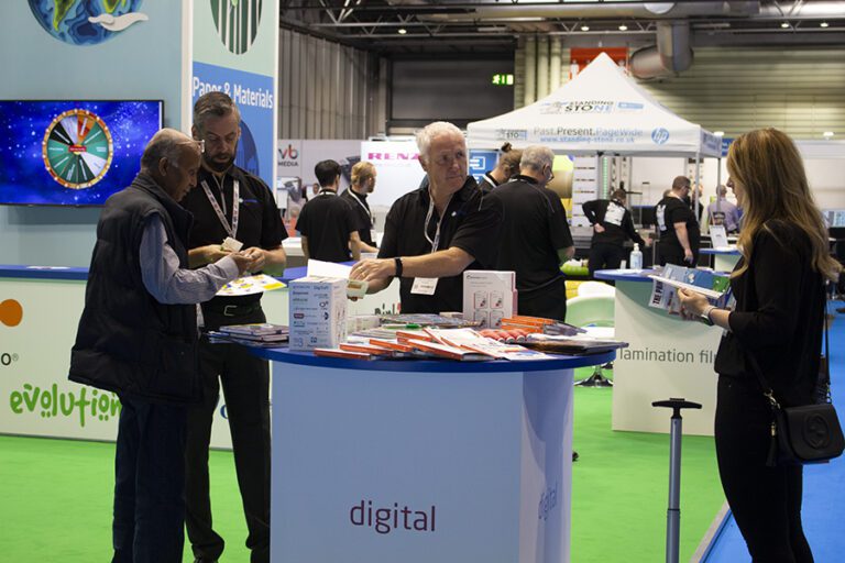 The Premier Paper stand at a UK trade show. The floor is green and there is a group of people wearing smart business clothes standing around a high table