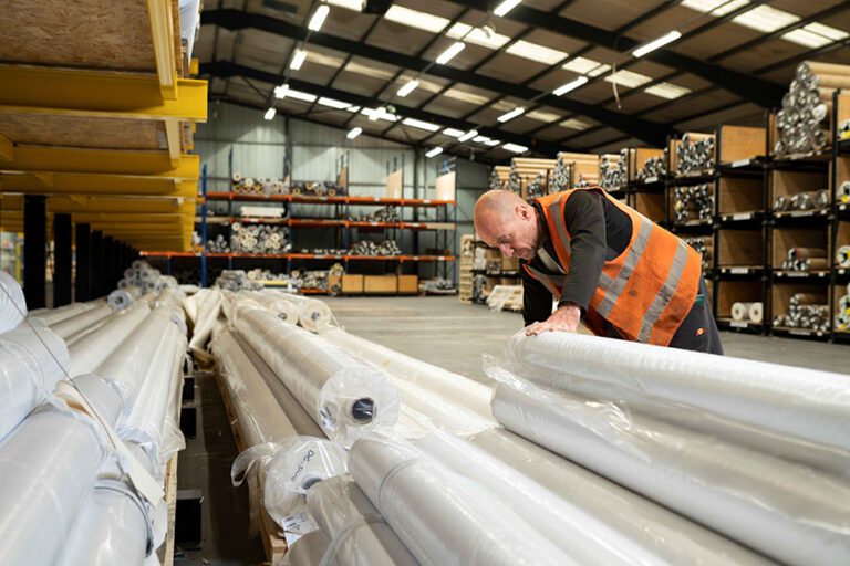 An image of inside Soyang's warehouse holding rolls of printable media. There is a warehouse worker on the right hand side wearing an orange hi-vis jacket and leaning over one of the piles of rolls to open it